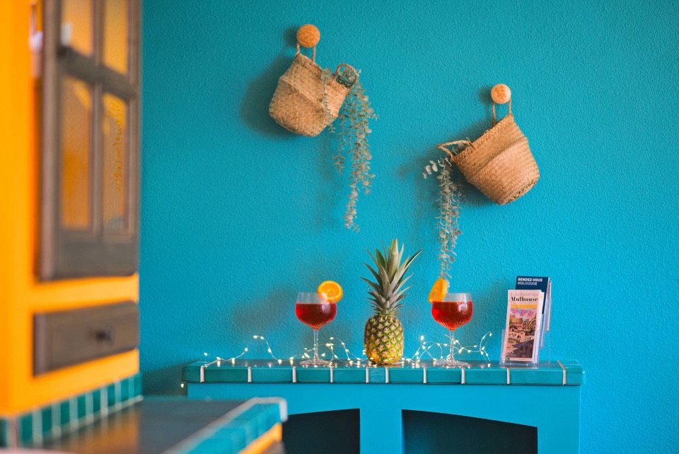 Console devant un mur turquoise avec paniers suspendus, deux spritz, un ananas et une guirlande lumineuse