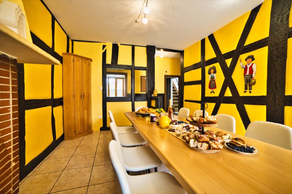 Dining room of an Alsatian holiday home with yellow walls and timber framing, long table set for breakfast
