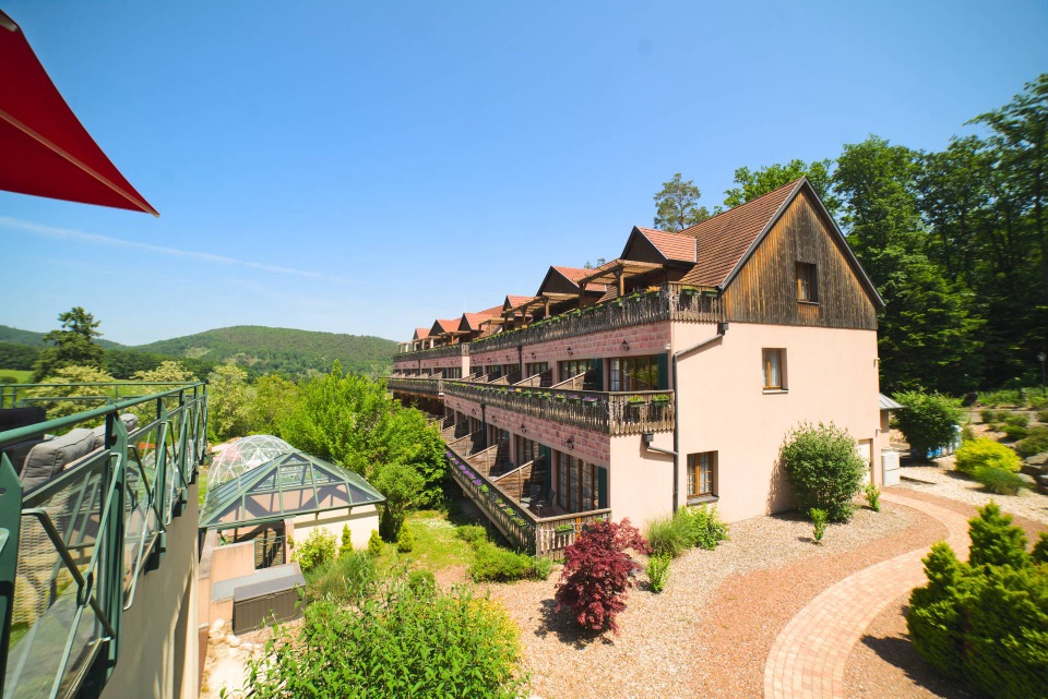 Hôtel de campagne alsacien aux balcons fleuris, collines boisées sous ciel bleu